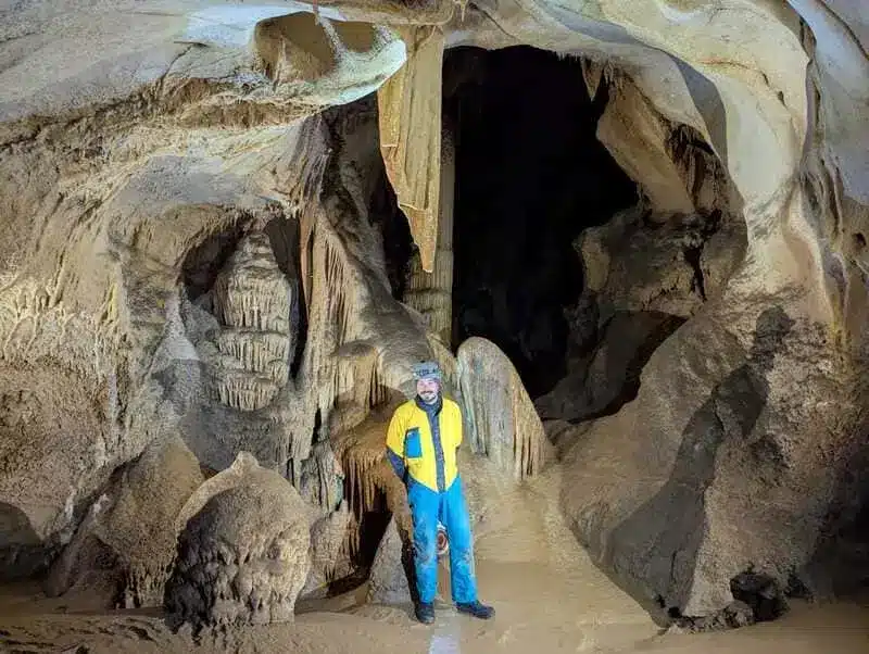 Une personne en tenue de spéléologie, prends la pose devant une stalagmite, dans une grotte des Hautes-Alpes
