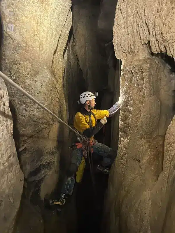 Iannis guide de spéléologie à Céüse descends sur une corde dans une grotte à l'aide d'un baudrier, sa lampe frontale éclaire la paroi de gauche