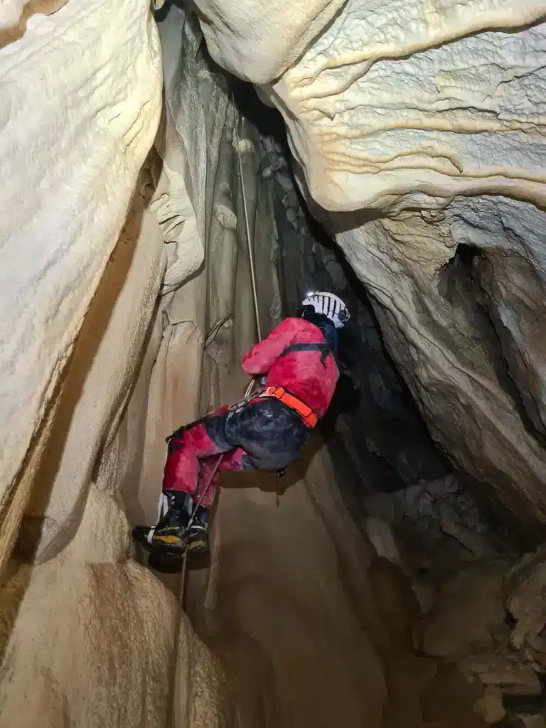 Vue de dessous d'un spéléologue dans une grotte naturelle qui remonte sur une corde à l'aide d'un baudrier