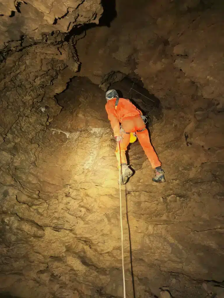 Un spéléologue en tenue de spéléologie de couleur orange descend un rappel un puits dans une grotte