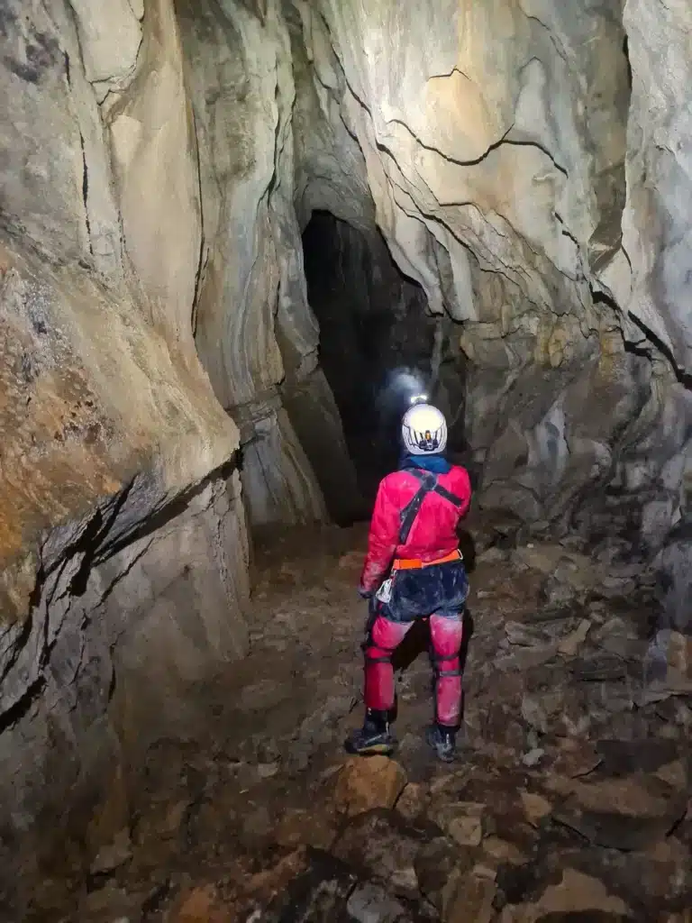 Une personne habillée en combinaison rouge, équipé d'un casque et d'une lampe frontale éclaire une grande galerie de la grotte de Céüse