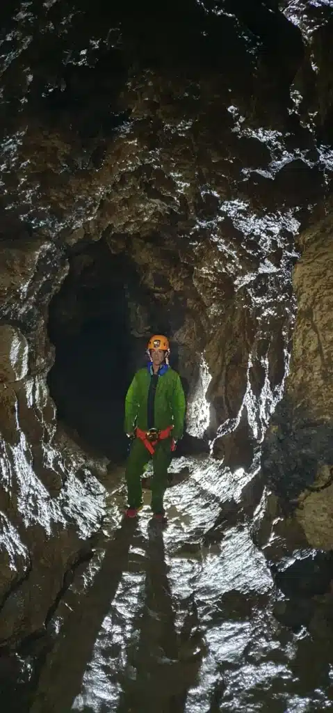Une personne équipée d'un casque d'une lampe frontale et d'une combinaison de spéléologie de couleur verte se tient debout dans une grande galerie de la grotte naturelle du Puits des Bans