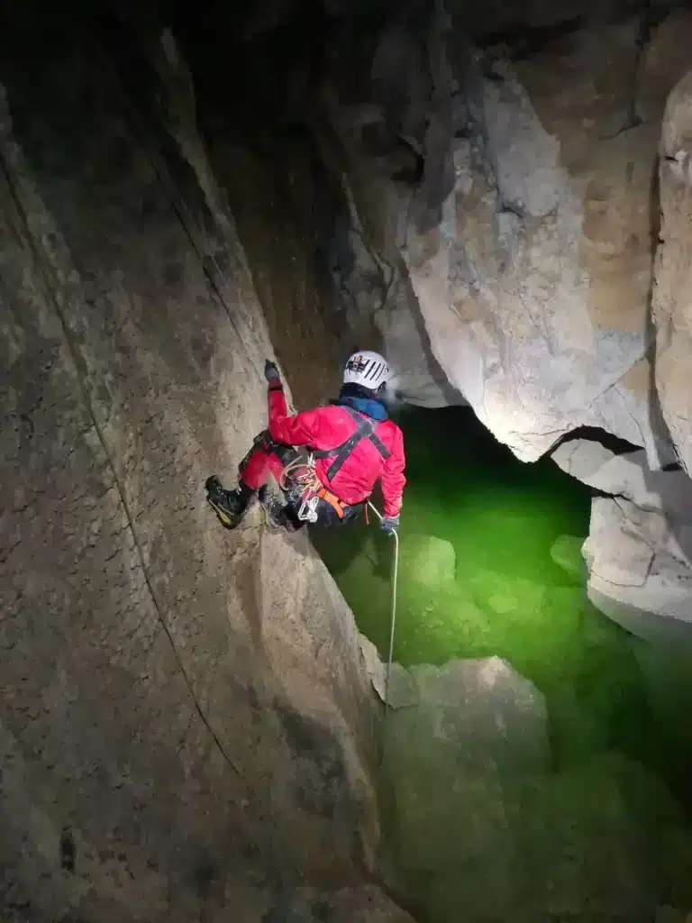 Une personne en tenue de spéléologie accrochée sur une corde avec un harnais de sécurité éclaire un lac souterrain avec son casque et sa lampe frontale