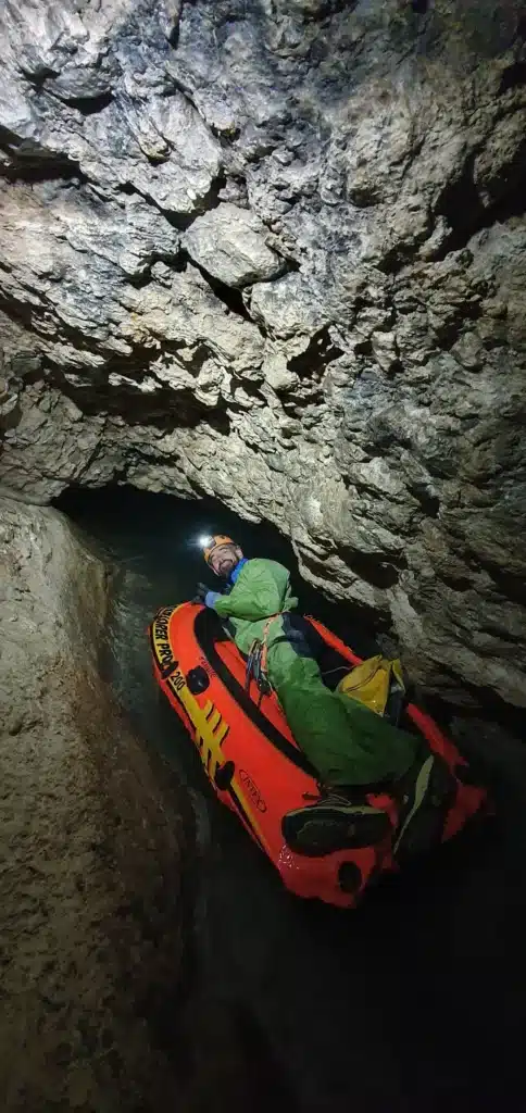 Une personne équipée d'un casque d'une lampe frontale et d'une combinaison de spéléologie de couleur verte est allongée sur un petit bateau gonflable pour franchir le petit lac d'entrée de la grotte du Puits des Bans
