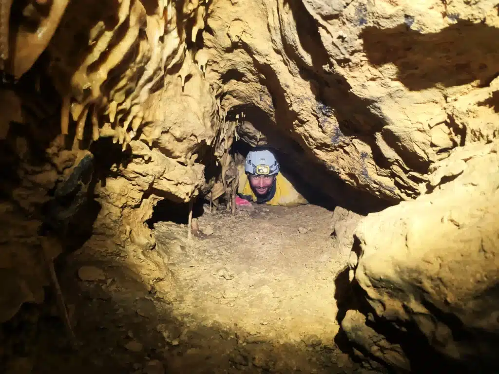 un spéléologue équipé d'un casque blanc, d'une lampe et d'une combinaison jaune sort d'un boyau dans lequel il doit ramper