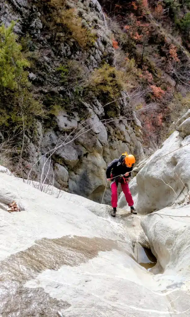 Une personne en équipement de canyoning, casque orange et pantalon rose, descend une cascade rocheuse à l'aide d'une corde à Orpierre.