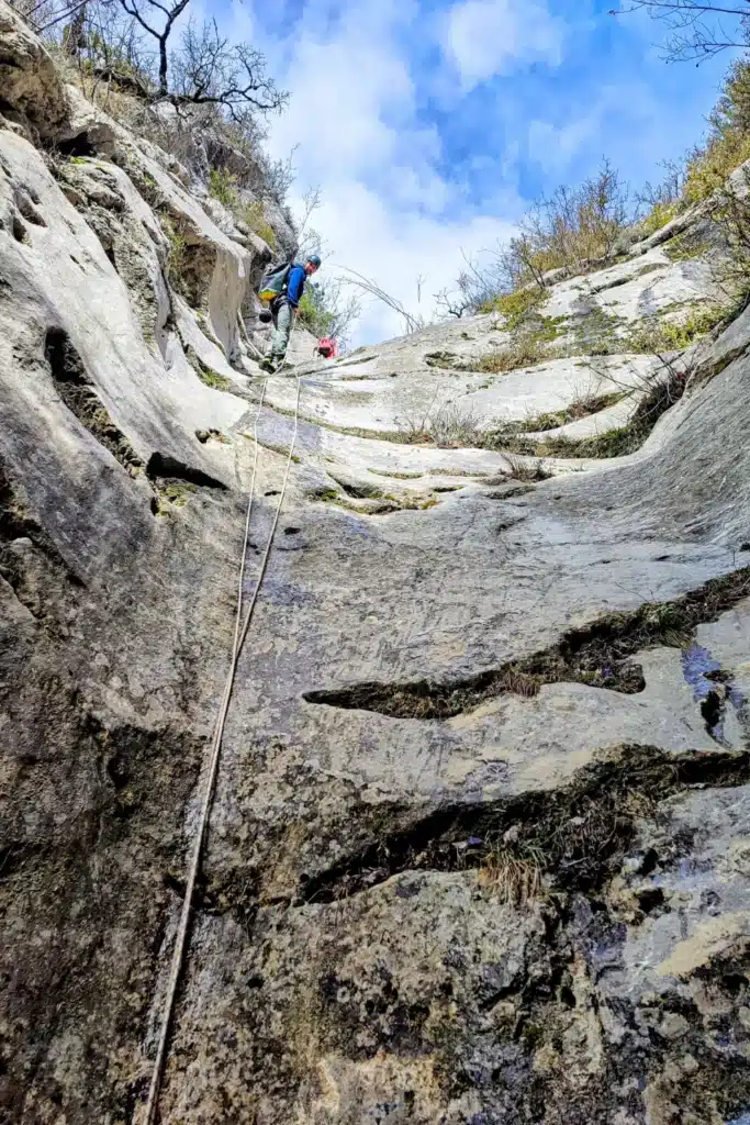 Un homme en combinaison de canyoning bleue descend en rappel sur une paroi rocheuse, le ciel bleu et les arbres dénudés en arrière-plan.