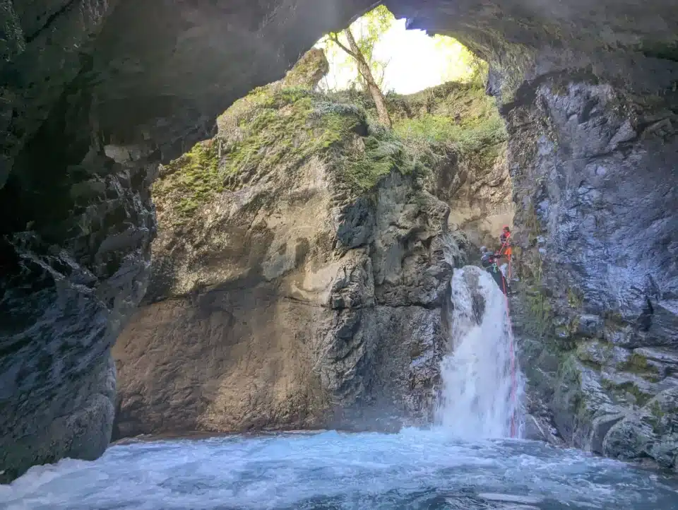 Une personne en combinaison fait du canyoning en descendant en rappel une cascade à l'intérieur d'une grotte rocheuse, avec de l'eau vive au premier plan.