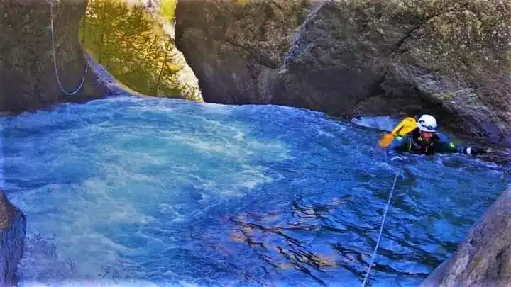 Une personne en tenue de canyoning avec un casque et un sac jaune se déplace dans une rivière d'eau bleue vive bordée de rochers.