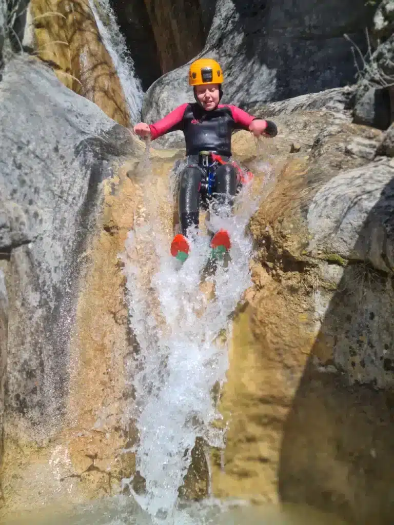Un enfant équipé d'un casque orange et d'une combinaison néoprène rouge glisse sur un toboggan naturel du canyoning des Marmites du diable