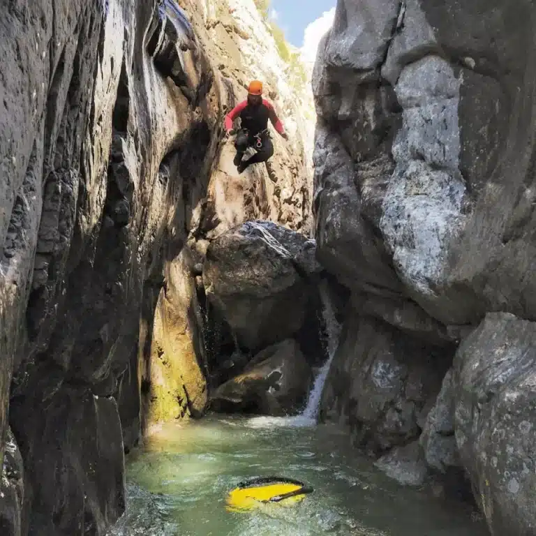 Un canyoniste en tenue de sport orange et noire saute au-dessus d'une piscine naturelle d'eau verte dans un canyon rocheux resserré.