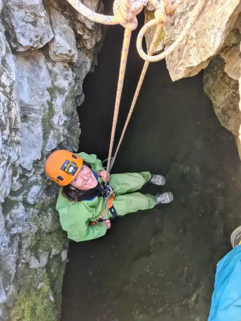 Une femme souriante en tenue de spéléologie verte et casque orange descend en rappel dans un Chourum du Dévoluy.