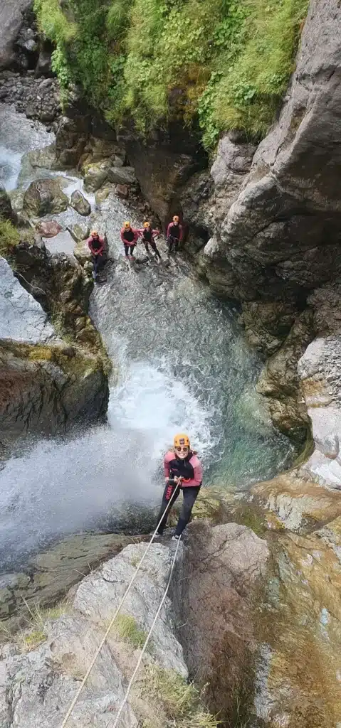 Une dame équipée d'un casque orange et d'une combinaison de canyoning rouge descend en rappel une cascade à Orcières Merlette.