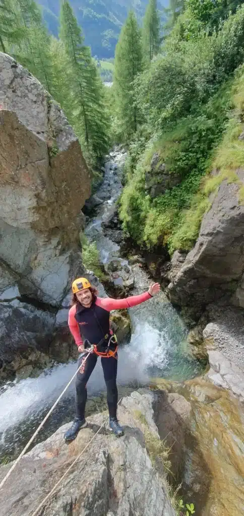 Un homme souriant en combinaison de canyoning et casque orange est prêt pour une descente, corde attachée, au-dessus d'une cascade dans un parcours de canyoning sportif à Orcières Merlette.