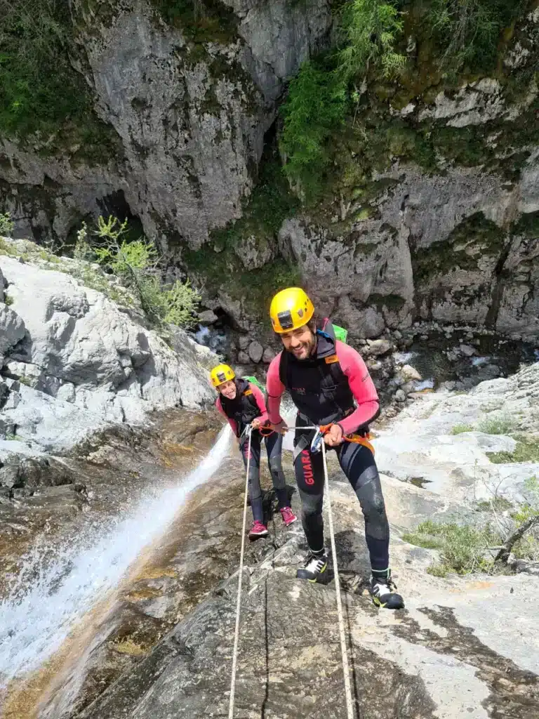 Un homme et une femme en équipement de canyoning descendent une cascade à Ancelle, protégés par des casques jaunes et retenus par des cordes.