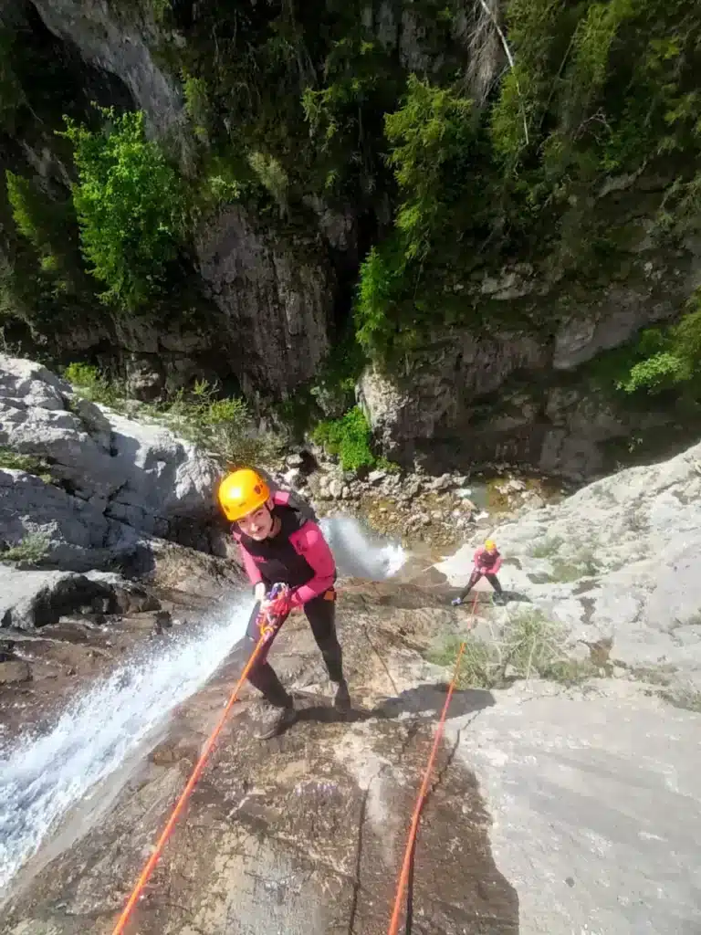 Un homme en tenue de canyoning rose et casque orange descend une cascade rocheuse à Ancelle, à l'aide d'une corde,