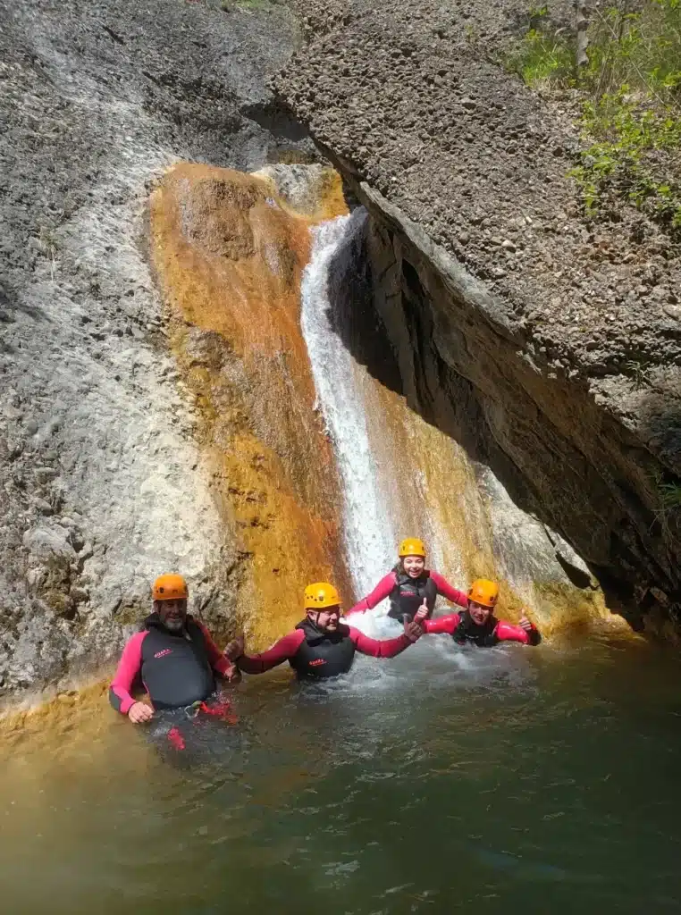Quatre personnes en combinaison néoprène et casque jaune posent dans un bassin au pied d'une cascade naturelle aux roches colorées.
