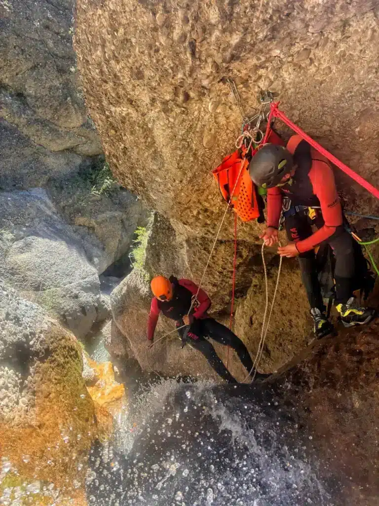 Deux personnes équipées pour le canyoning descendent en rappel une cascade rocheuse, l'une tenant la corde au-dessus de l'eau tumultueuse. Ils sont dans le massif du Dévoluy.