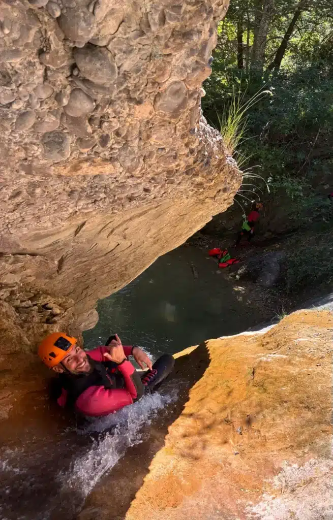 Un homme en combinaison et casque orange fait du canyoning dans le Dévoluy, glissant sur un toboggan naturel vers une rivière claire. D'autres canyonistes en arrière-plan.