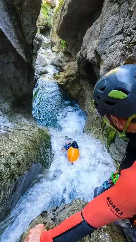 Une personne en combinaison orange et casque bleu descend un toboggan naturel dans une cascade dans des gorges rocheuses étroites. Une autre personne est juste derrière, dans la chute d'eau du canyon d'Orcières Merlette.