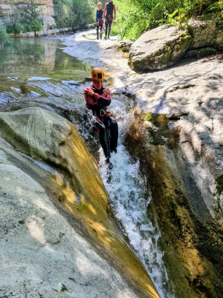 Une personne en combinaison et casque de canyoning glisse sur une cascade dans un canyoning à Rabou, avec de l'eau qui bouillonne autour d'elle.