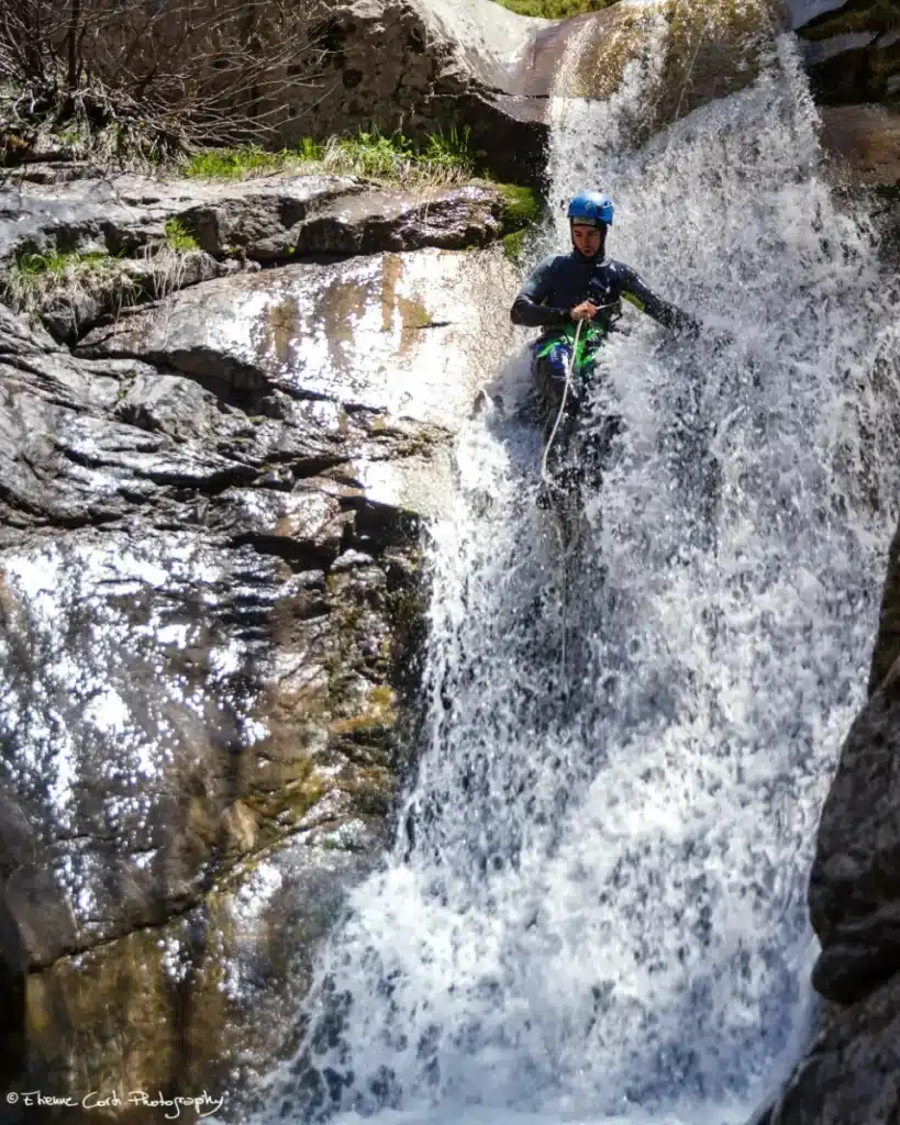 Un homme en tenue de canyoning, casque bleu, descend une cascade sur une corde, entouré d'éclaboussures d'eau, dans le canyon de rouanette à Ancelle.