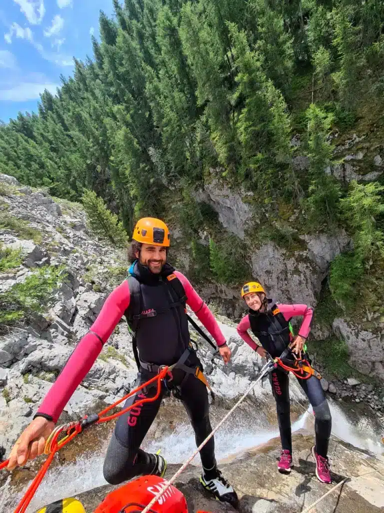 Un homme et une femme souriants en combinaison de canyoning et casque orange, préparés pour descendre le cascade de Peyron Roux à Ancelle, entourée de végétation dense.