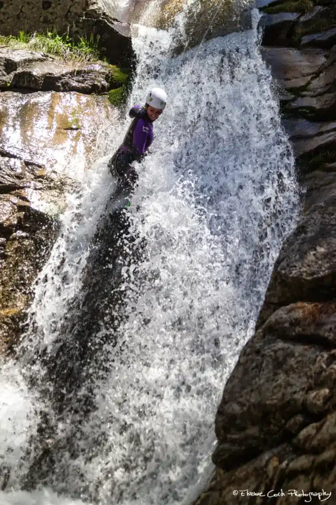 Une femme en combinaison violette et casque blanc descend une cascade lors d'une activité de canyoning à Ancelle.