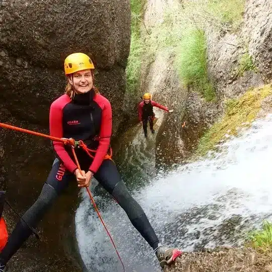 Une femme souriante en combinaison rouge et noire et casque jaune descend en rappel le long d'une cascade en canyoning du Dévoluy. Une autre personne est visible plus bas.