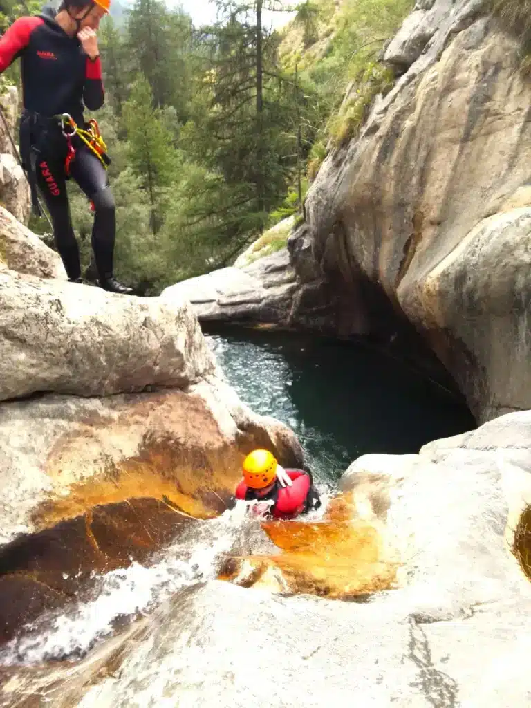 Une personne en combinaison néoprène et casque orange glisse sur un toboggan naturel dans un canyon verdoyant à Ancelle.