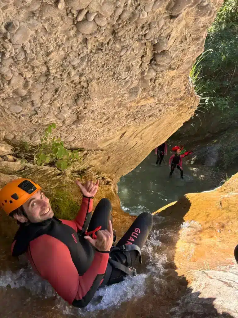 Un homme souriant en combinaison, casque orange, faisant un signe "shaka" dans un toboggan naturel du canyon du Dévoluy avec d'autres canyonistes en arrière plan.
