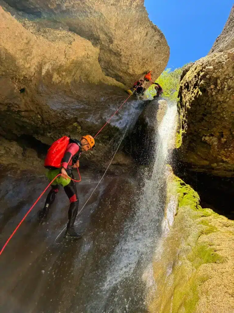 Canyoning Dévoluy : un homme descend une cascade en rappel, casque orange, sac à dos rouge, combinaison noire et verte.