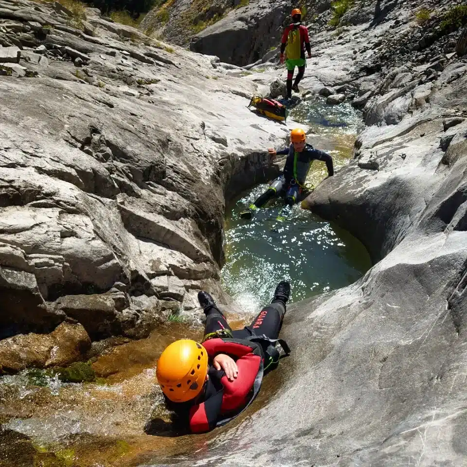 Trois personnes équipées pour faire du canyoning descendent le canyon de Peyron Roux à Ancelle, entre des rochers, une personne glisse sur un toboggan naturel, les deux autres personnes le regarde.