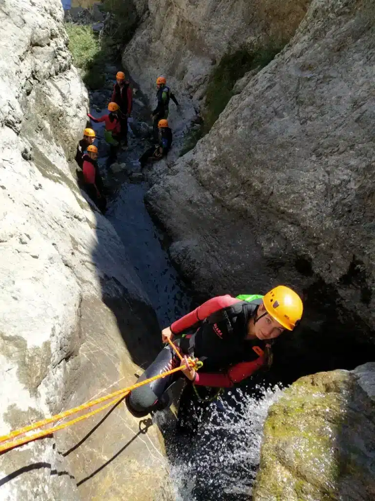 Un groupe de personnes en combinaison néoprène et casque s'engage dans le canyon de Peyron Roux à ancelle, l'une d'elles descend en rappel avec une corde orange.