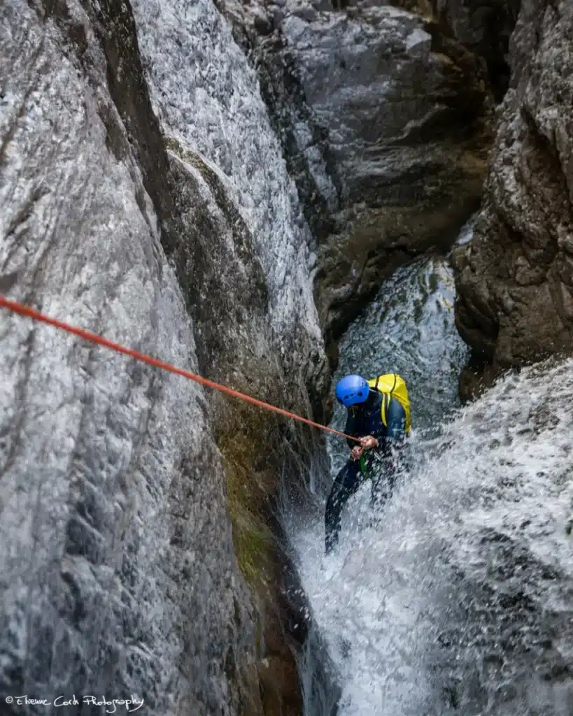 Personne en combinaison bleue et casque bleu descendant en rappel une cascade d'eau vive dans le canyon de Rouanette intégrale à Ancelle qui est étroit, avec un sac à dos jaune.