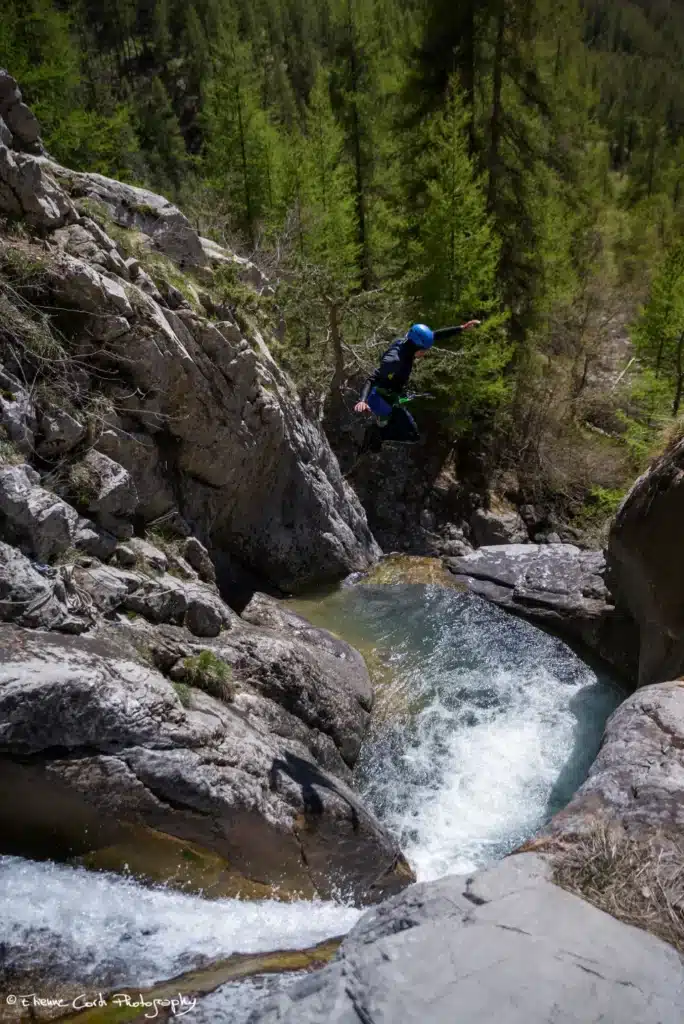 Un canyoniste en combinaison bleue et casque bleu saute dans une cascade rocheuse entourée de forêt à Ancelle.