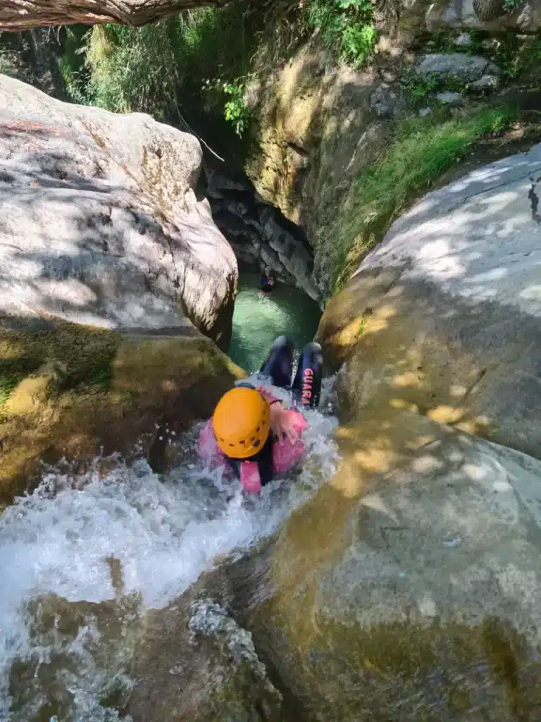 Personne en combinaison rose et casque jaune descendant le canyon de Rabou sur un toboggan naturel, avec une autre personne dans l'eau en arrière-plan.