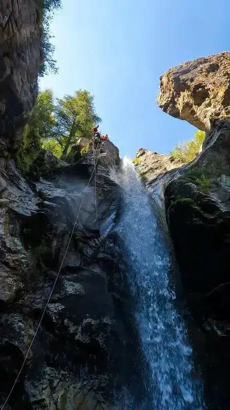 Des personnes font du canyoning à Orcières Merlette, descendant en rappel une haute cascade avec de l'eau par une journée ensoleillée.