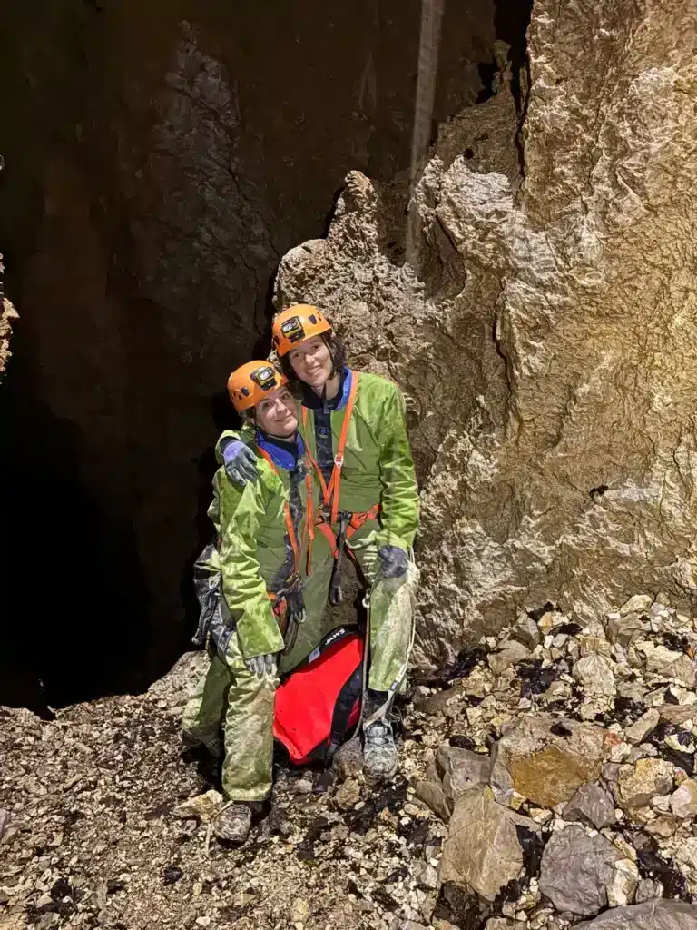 Deux personnes souriantes, équipées de casques, combinaisons vertes et baudriers, posent dans une grotte qui s'appelle le Chourum Carmarguier situé dans le massif du Dévoluy.