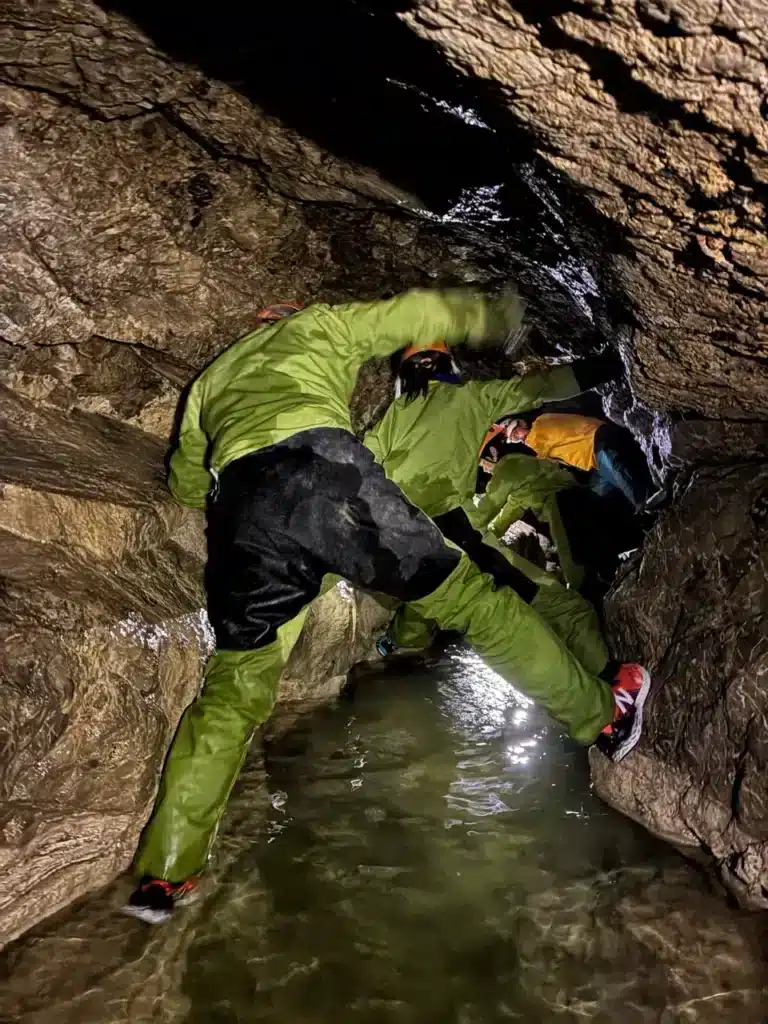 Trois explorateurs en combinaison verte se faufilent dans un passage étroit d'une grotte inondée du massif du Dévoluy, au dessus de l'eau et les rochers mouillés.