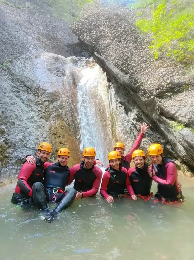 Un groupe de huit personnes souriant, portant des combinaisons de canyoning et des casques, se tient dans une eau peu profonde devant un toboggan naturel dans une rivière.