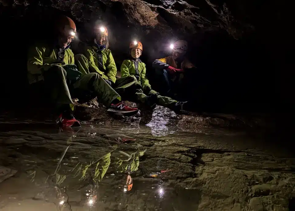 Quatre personnes, équipées de casques avec lampes frontales, sont assises dans une grotte du Dévoluy, avec leurs reflets dans l'eau au premier plan.