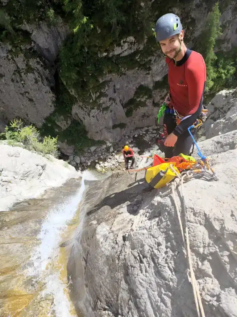 Deux canyonistes équipés de casques et de combinaisons se préparent à descendre une cascade avec de l'eau, l'un en premier plan est le guide de canyoning du village d'Ancelle, l'autre est plus loin près du point de départ.