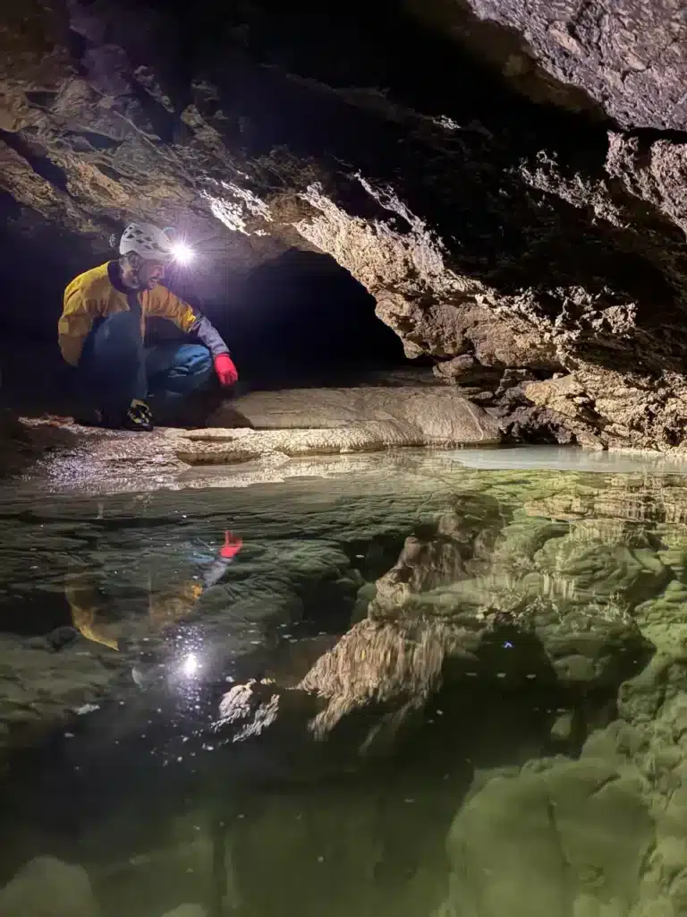 Iannis Wypyszynski est un guide de spéléologie éclairé par sa lampe frontale accroupi près d'une étendue d'eau dans une grotte du Dévoluy.
