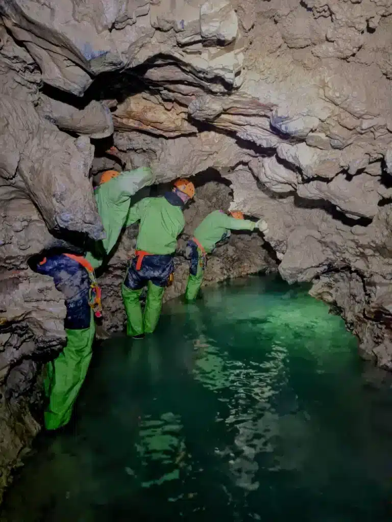 Trois spéléologues équipés de combinaisons vertes et de casques évoluant dans une grotte près d'un plan d'eau à la couleur verte intense.