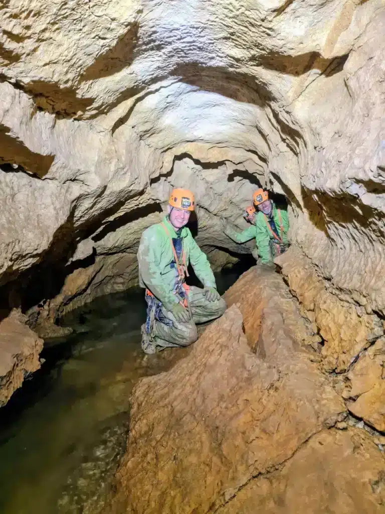 Trois explorateurs en combinaison verte et casque orange souriant à l'intérieur de la grotte de la Fountarrasse, avec une rivière souterraine à leurs pieds.