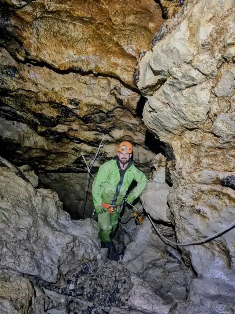 Un homme portant un casque orange et une combinaison verte est debout dans une grotte du Dévoluy qui se nome le Puits des Bans. Il est équipé pour la spéléologie.
