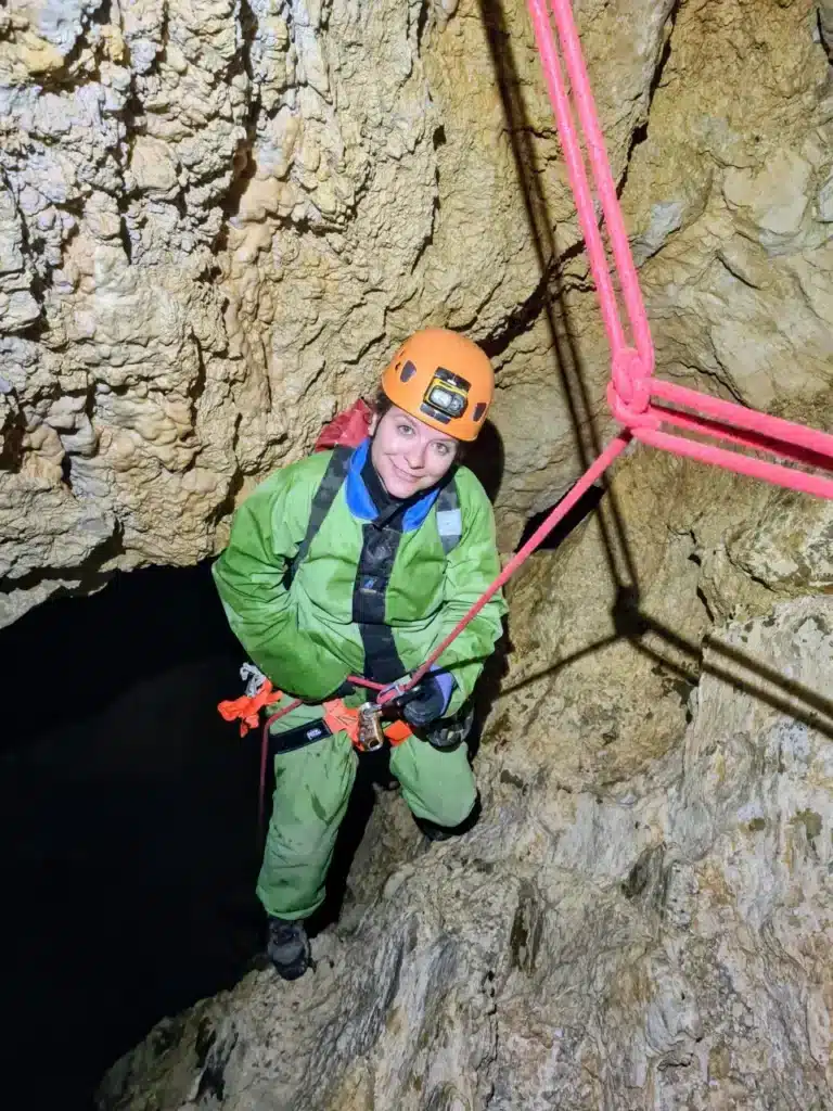 Femme souriante en casque orange et tenue de spéléologie verte, équipée pour la descente en rappel dans le Chourum Camarguier qui est une grotte du Dévoluy.