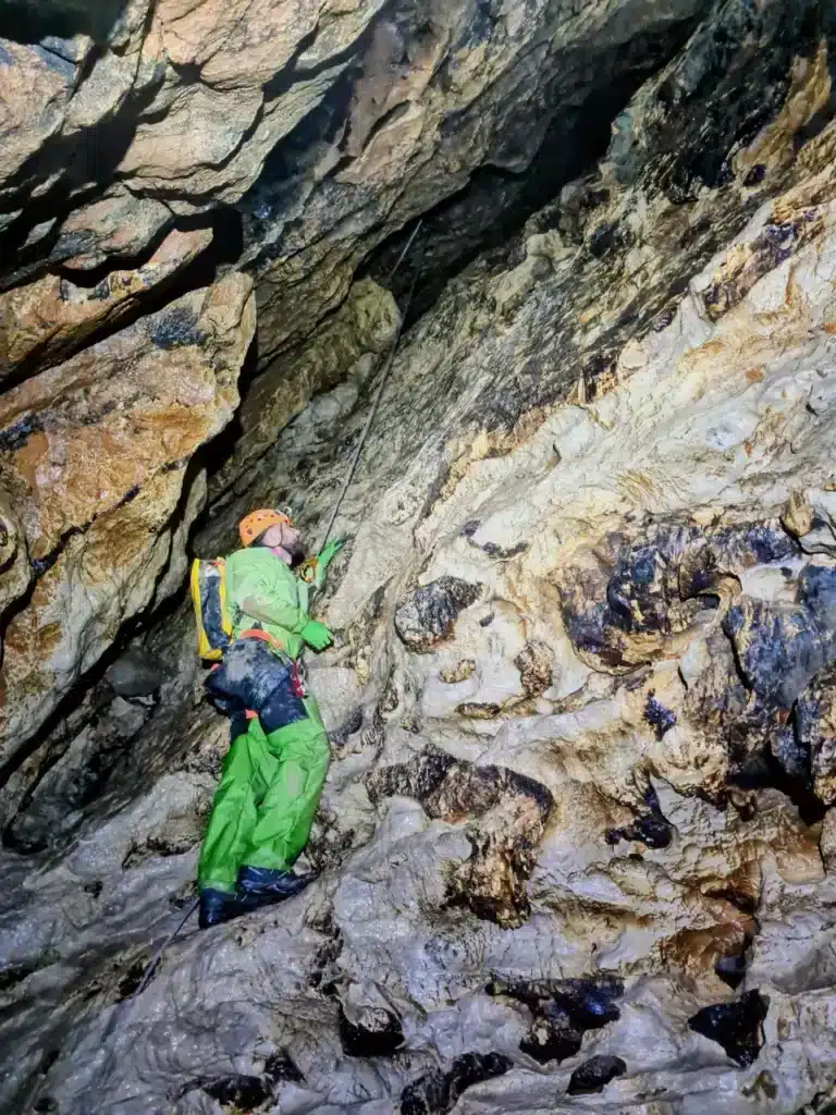 Un spéléologue en combinaison verte et casque orange progresse le long d'une paroi rocheuse humide à l'aide d'une corde dans le Puits des Bans.