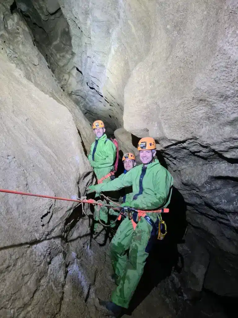 Trois spéléologues en combinaison verte et casque orange posent dans une étroite cavité rocheuse de la vallée du Buëch, une corde rouge passant devant eux.
